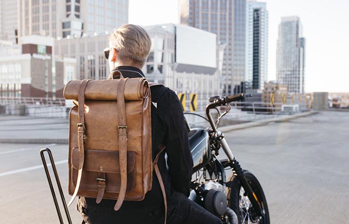 A man on a motorbike on a city street with a Leather Rolltop Backpack by Johnny Fly Co. on his back, rear view.