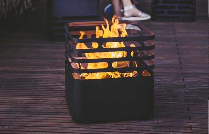 Höfats CUBE Fire Pit, with fire burning inside, on a wooden floor with a person's feet in the background.