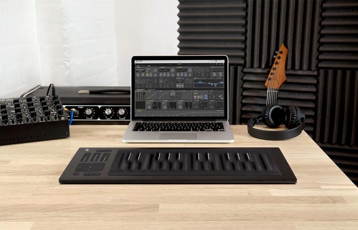 Roli Seaboard Rise on a wooden table with a laptop and headphones and a guitar and music production equipment in the background.