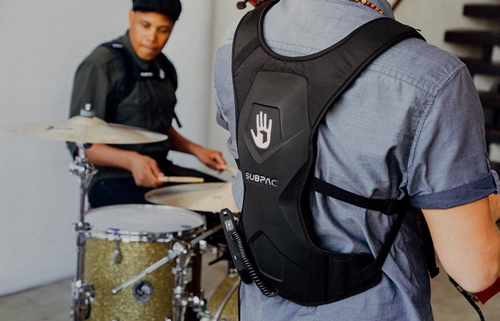 SubPac M2 worn by a man in a grey tee shirt, back view, tilted, and a man behind the drums.