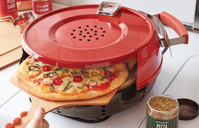 Pizzacraft Stovetop Pizza Oven on a gas stove with a pizza on a wooden pizza peal halfway in.