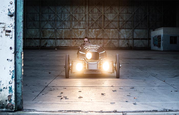 A Guy Driving Morgan EV3 Electric Car With The Lights Turned On