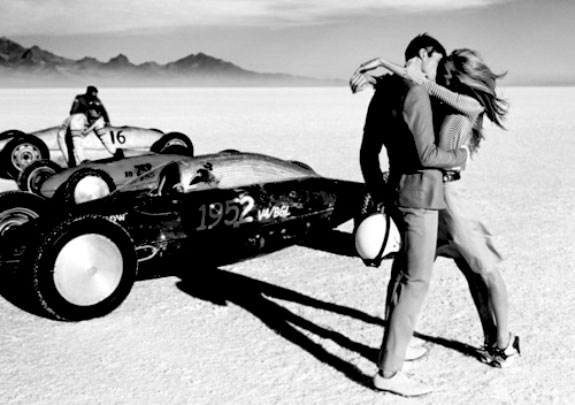 Black and white photo two people kissing in the dessert next to the racer car