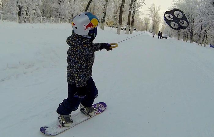 A little boy on a snowboard with a helmet being pulled across the snow by a drone.