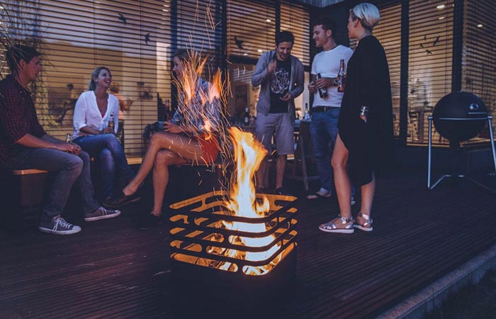 Höfats CUBE Fire Pit with fire lit inside, on a terrace, and people socializing in the background.