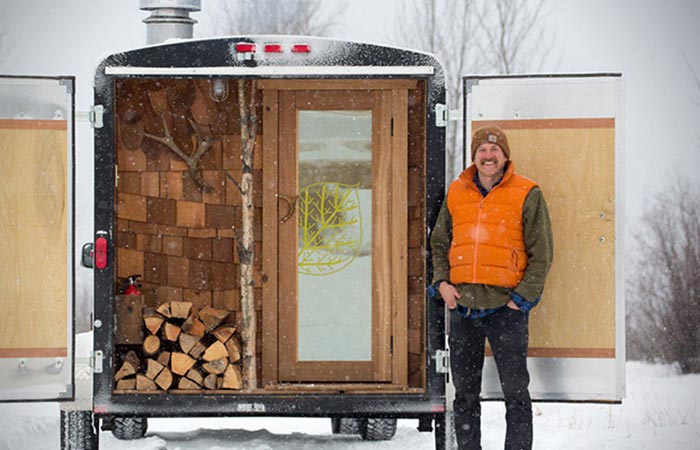 A man standing by the sauna on wheels. 